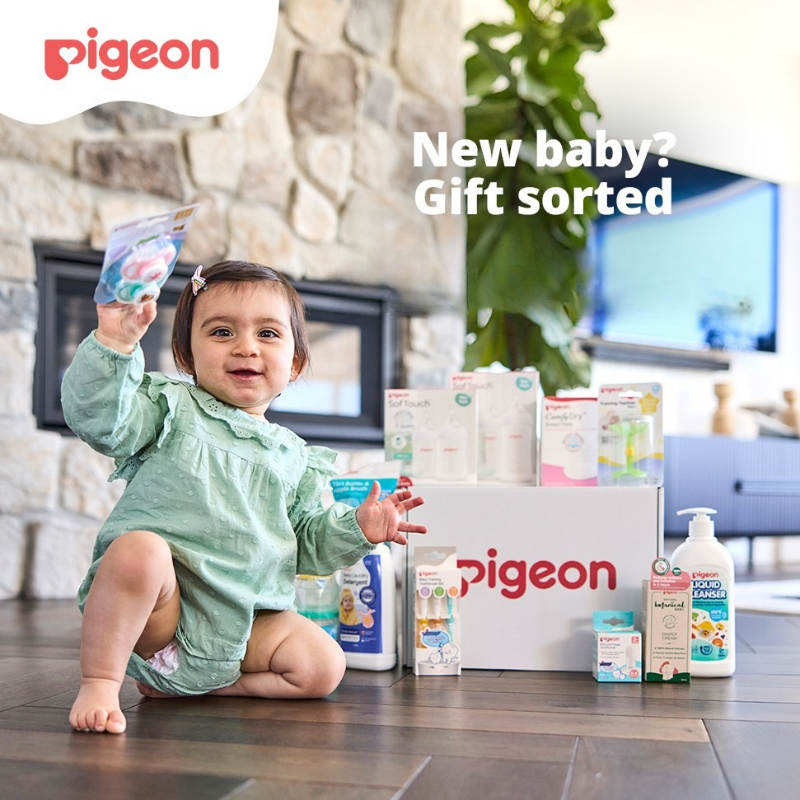 Baby girl sitting on the floor holding up a Pigeon dummies twin pack, with the Pigeon Gift Box and its baby essentials beside her
