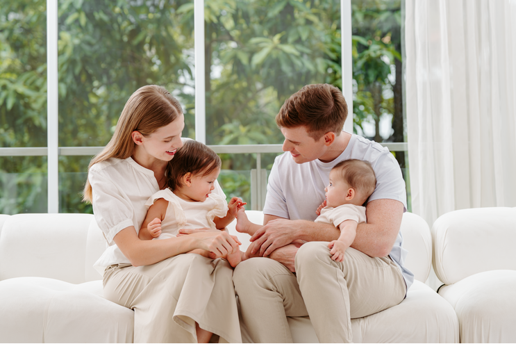 Family of four sitting on a white couch in a bright room with large windows.